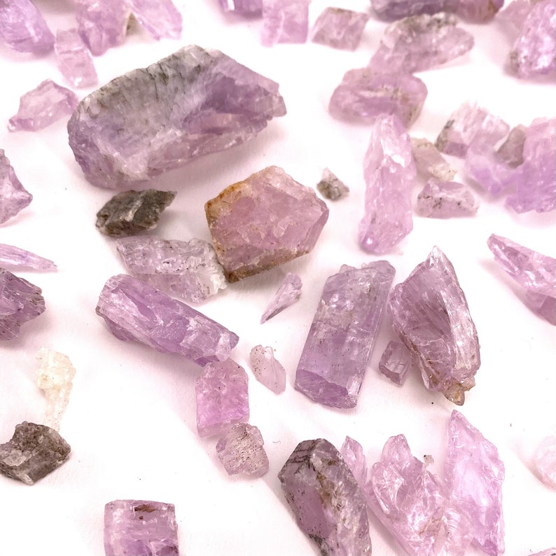 K&ouml;nnte beinhalten: Eine Sammlung von rosa und violetten Kristallen, einige mit einem leichten Graustich, verstreut auf einer wei&szlig;en Oberfl&auml;che. Die Kristalle sind in verschiedenen Formen und Gr&ouml;&szlig;en, einige sind lang und d&uuml;nn, w&auml;hrend andere runder sind.