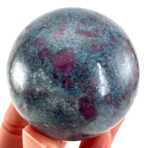 Könnte beinhalten: Eine polierte Edelstein-Kugel, überwiegend blau-grau mit auffälligen, unregelmäßigen Flecken in tiefem Magenta. Die Oberfläche des Steins ist glatt und reflektierend und zeigt die natürlichen Muster und Farben des Minerals. Die Kugel wird in einer Hand gehalten.