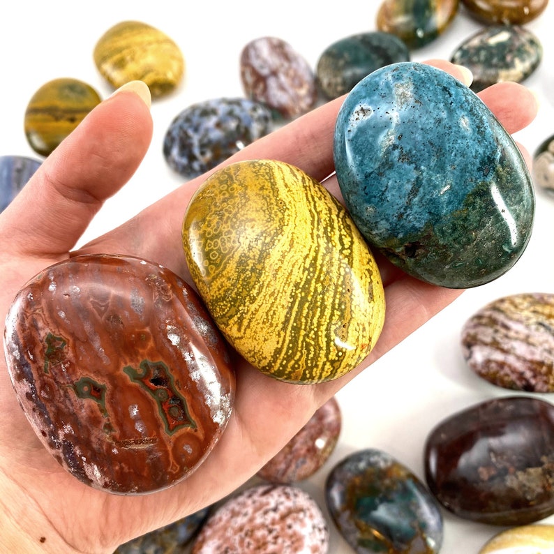 May include: A selection of polished stones in diverse colours and patterns. The stones feature shades of red, yellow, blue, and brown. Some stones display unique veining and speckled designs. The stones are smooth and oval-shaped, ideal for collecting or display.