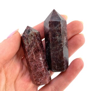 May include: Two red and brown crystal points, also known as red phantom quartz, are held in a hand. The points are about 2 inches long and have a smooth, polished finish.