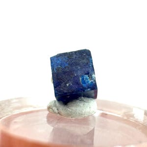 May include: A dark blue, cube-shaped mineral specimen with a rough texture. The stone has patches of lighter blue and sits on a small, off-white base. The background is a soft pink and white.
