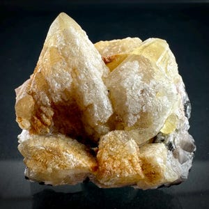 Puede incluir: Un grupo de minerales cristalinos de color amarillo pálido y blanco. Los cristales tienen una superficie rugosa y texturizada y están dispuestos en una formación en capas. La formación mineral tiene un pico afilado y puntiagudo.