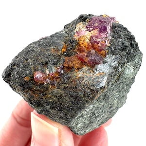 May include: A rough, dark gray rock with embedded, vibrant pink and red crystals. The rock has a textured surface with patches of orange and brown. The crystals are irregularly shaped and appear to be partially exposed.