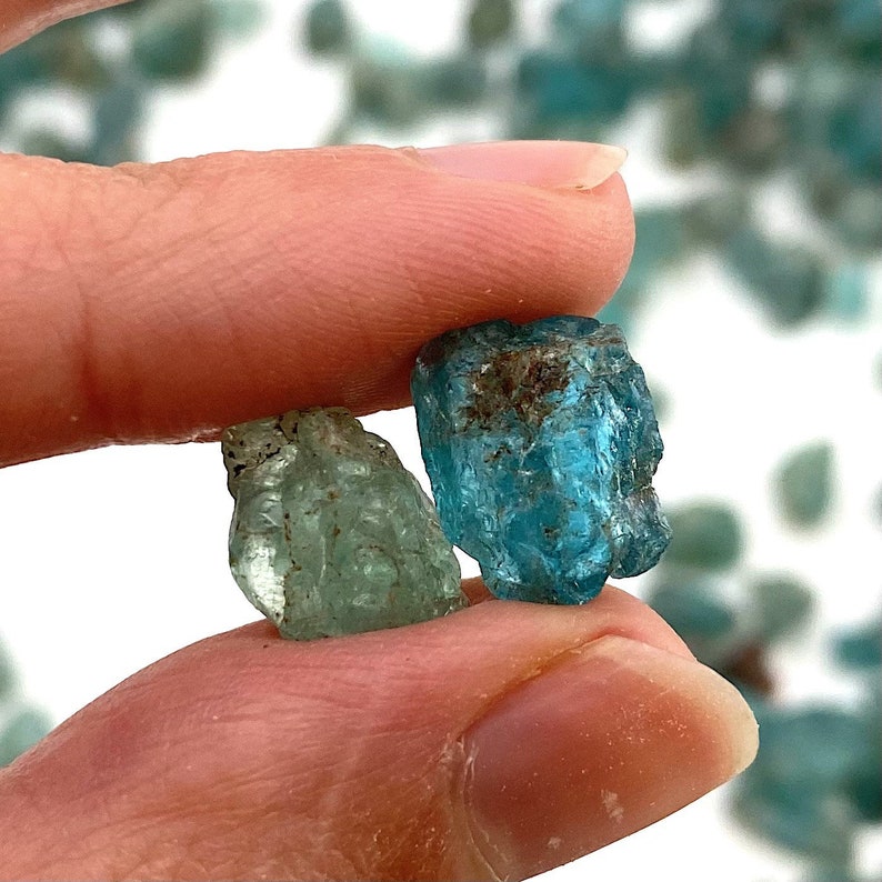 May include: Two rough, faceted gemstones, one blue and one green, are held in a hand. The blue gemstone is larger and has a more rounded shape, while the green gemstone is smaller and more elongated. Both gemstones have a translucent appearance.