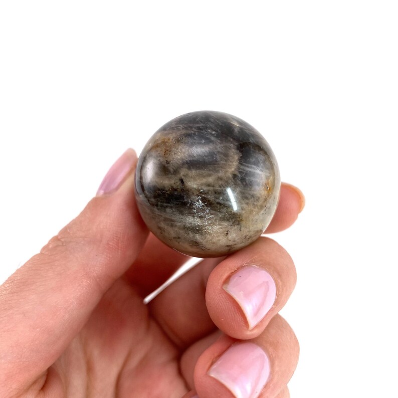 May include: A smooth, polished black and grey sunstone sphere. The sphere is held in a hand with pink nail polish.