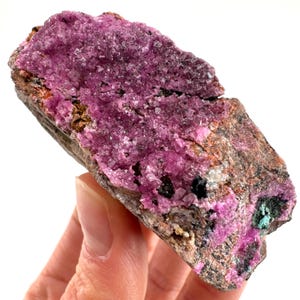 May include: A pink and purple cobalt calcite specimen with a rough, natural texture. The calcite is embedded in a matrix of brown and grey rock.