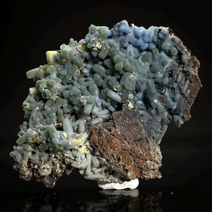 May include: A mineral specimen featuring a cluster of pale blue-green, cylindrical crystals growing from a dark, textured, brownish-black matrix. The crystalline structure displays a dense, branching formation with small, reflective metallic inclusions.