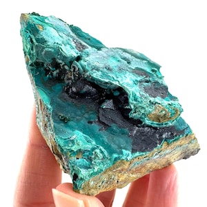 May include: A natural malachite specimen, displaying a striking teal and black colour scheme. The mineral has a rough, textured surface with areas of deep green and dark, almost black, inclusions. The edges show a golden-brown hue.
