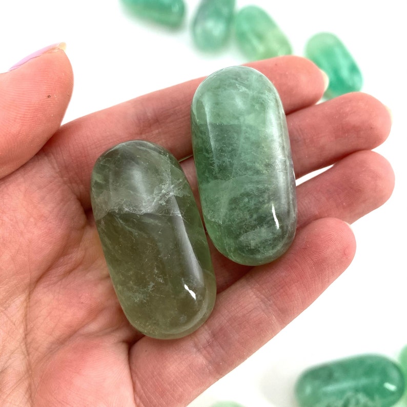 May include: Two green fluorite tumbled stones, also known as polished stones, in a hand. The stones are smooth and have a light green color.