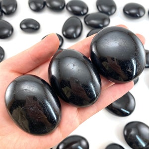 May include: Three smooth, polished, oval-shaped black stones held in a palm, with several other identical black stones scattered in the background. The stones have a glossy, reflective surface with subtle natural surface textures visible.