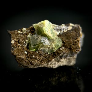 Pode incluir: Uma imagem de perto de um espécime mineral com um grupo de cristais translúcidos verde claro. Os cristais estão dispostos sobre um fundo de rocha matriz castanha áspera. O mineral tem uma superfície reflexiva e o fundo é preto.