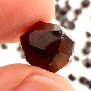 May include: A deep red, faceted gemstone with a natural, slightly cloudy appearance. The gemstone is a crystal with many facets and is held in a hand.