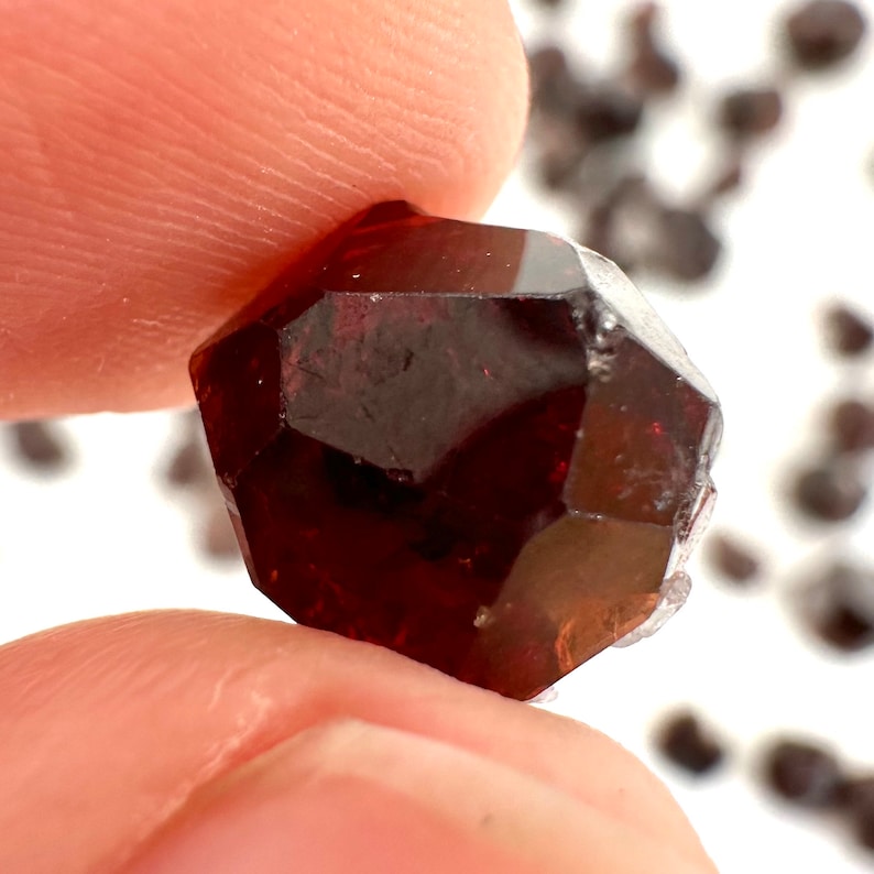 May include: A deep red, faceted gemstone with a natural, slightly cloudy appearance. The gemstone is a crystal with many facets and is held in a hand.