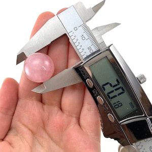 May include: A light pink rose quartz sphere, approximately 20 mm in diameter, is measured with a digital caliper. The caliper displays a measurement of 20.18 mm. The sphere is held against a white background.