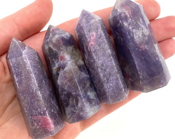 May include: Four polished, purple-hued crystal towers with pink inclusions are held in a hand against a white background. The crystals have a faceted, tapered shape, and vary slightly in color and pattern. The crystals are a natural, decorative item.