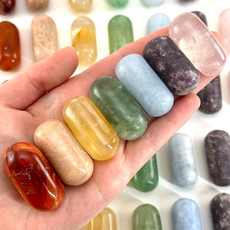 May include: A set of eight smooth, polished, tumbled stones in various colours, including red, orange, yellow, green, blue, purple, and pink. The stones are arranged in a rainbow pattern and are held in a hand.