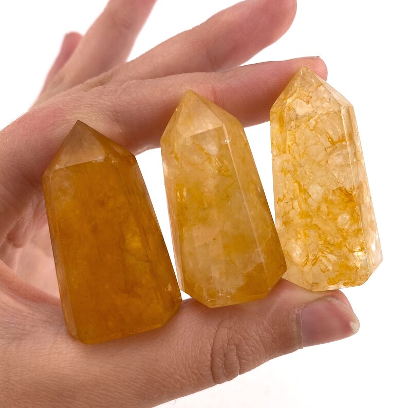 May include: Three golden-yellow crystal points with varying shades and textures are held in a hand. The crystals have a polished, faceted appearance, with a tapered shape and a flat base. The crystals are a natural, earthy product.