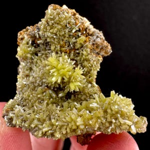 May include: A close-up of a mineral specimen featuring clusters of small, light yellow-green crystals. The crystals are needle-like and radiate outwards from a central point, creating a starburst effect. The base is a mix of brown and tan.