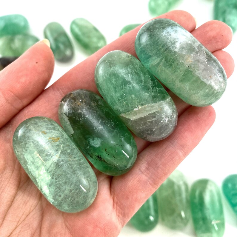 May include: A collection of smooth, green fluorite tumbled stones. The stones are oval shaped and have a translucent, glassy appearance.
