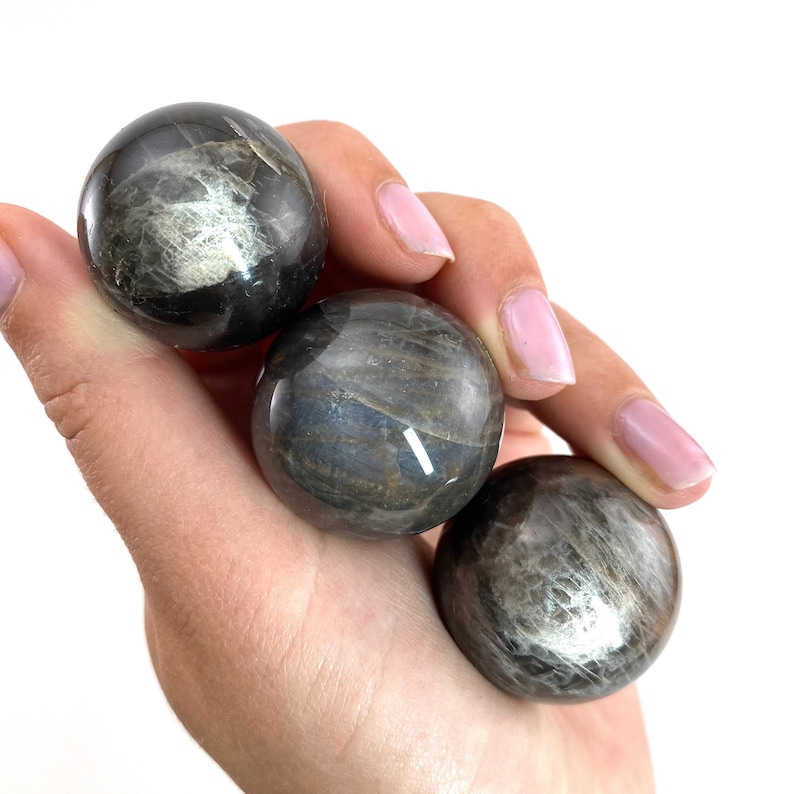 May include: Three black obsidian spheres with a silvery sheen, held in a hand. The spheres are smooth and polished, with a natural, earthy look.