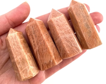 May include: Four orange sunstone crystal points, also known as sunstone towers, are arranged in a row in a hand. The points are polished and have a smooth, glassy finish.