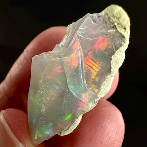 May include: A raw, iridescent opal gemstone with a clear, crystalline structure. The opal displays a vibrant play-of-color, with flashes of red, orange, yellow, and green. The stone has a rough, natural exterior and is held in a hand.