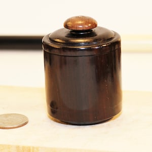 May include: A small, dark brown wooden jar with a lid. The lid has a round knob on top.