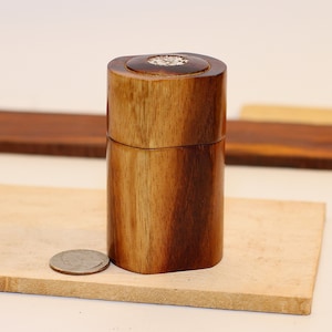 May include: A small, round, wooden box with a decorative top. The box is made of dark wood with a light brown grain. The box is sitting on a light brown surface.