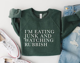 May include: A dark green sweatshirt with the text "I'm eating junk and watching rubbish" in white lettering. The sweatshirt is paired with light blue jeans and white sneakers.