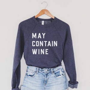 Puede incluir: Una sudadera azul marino con el texto "MAY CONTAIN WINE" impreso en blanco en la parte delantera. La sudadera se est&aacute; modelando con unos pantalones cortos de mezclilla azul claro.