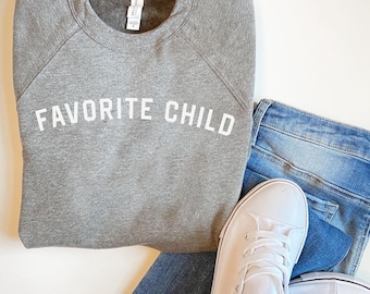 May include: A gray sweatshirt with the text "FAVORITE CHILD" in white lettering. The sweatshirt is paired with blue jeans and white sneakers.