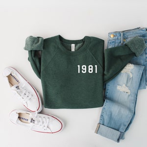 May include: A green sweatshirt with the year 1981 printed in white on the front. The sweatshirt is paired with light blue jeans and white sneakers.