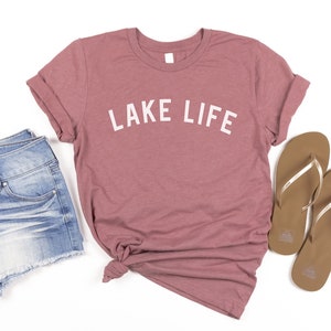 May include: A mauve t-shirt with the text "LAKE LIFE" in white lettering. The t-shirt is paired with denim shorts and brown flip-flops.