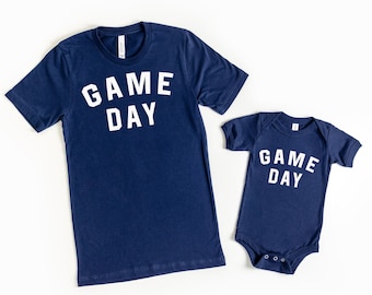 Puede incluir: Dos camisas azul marino con el texto "GAME DAY" en blanco. La camisa más grande es una camiseta y la camisa más pequeña es un body.