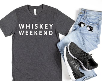 May include: A dark grey t-shirt with the text "WHISKEY WEEKEND" printed in white on the front. The shirt is paired with blue jeans and black sneakers.