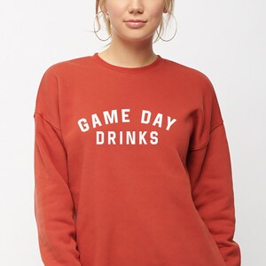 K&ouml;nnte beinhalten: Ein rostfarbener Pullover mit dem Aufdruck "GAME DAY DRINKS" in Wei&szlig;.