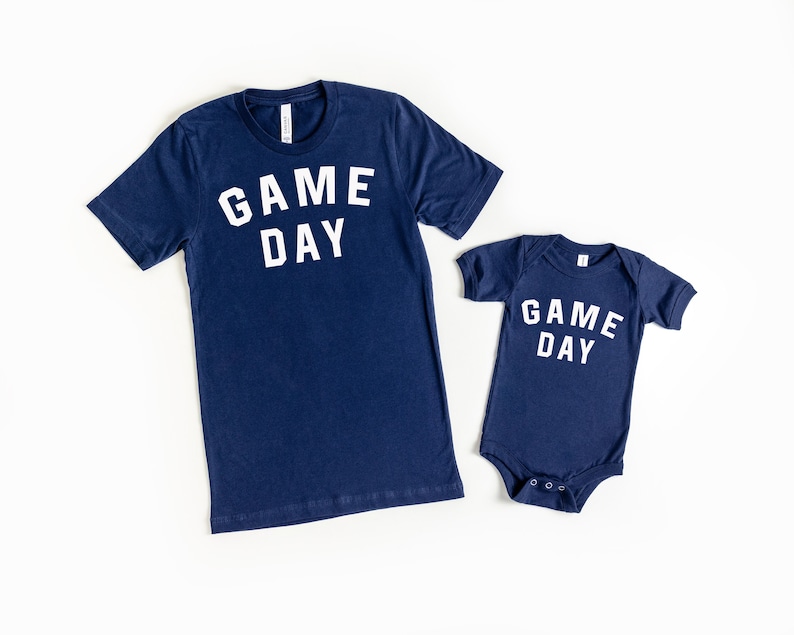 K&ouml;nnte beinhalten: Zwei marineblaue Shirts mit dem Text "GAME DAY" in Wei&szlig;. Das gr&ouml;&szlig;ere Shirt ist ein T-Shirt und das kleinere Shirt ist ein Body.