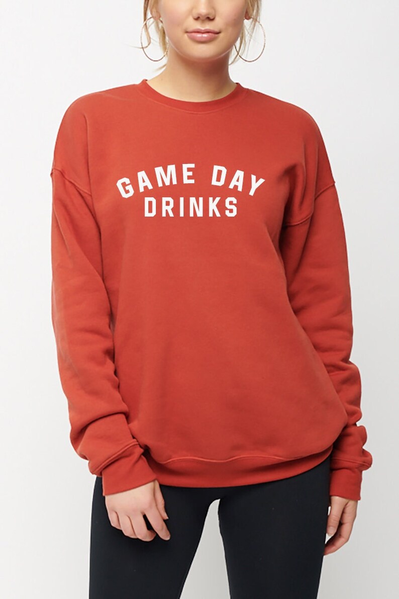 K&ouml;nnte beinhalten: Ein rostfarbener Pullover mit dem Aufdruck "GAME DAY DRINKS" in Wei&szlig;.