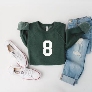 May include: An overhead shot of a casual outfit. A dark green sweatshirt with the number 8 in white is paired with light blue ripped jeans and white sneakers. The sneakers have red accents.