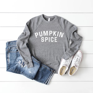 May include: A gray sweatshirt with the text "Pumpkin Spice" in white letters. The sweatshirt is paired with blue jeans and white sneakers.