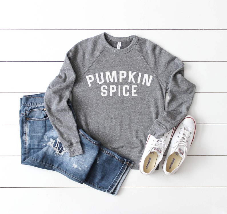 May include: A gray sweatshirt with the text "Pumpkin Spice" in white letters. The sweatshirt is paired with blue jeans and white sneakers.