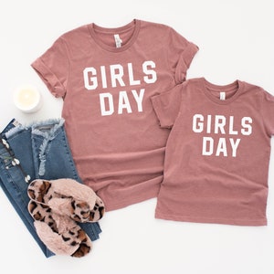 May include: Two t-shirts with the text "Girls Day" in white letters on a dusty rose background.