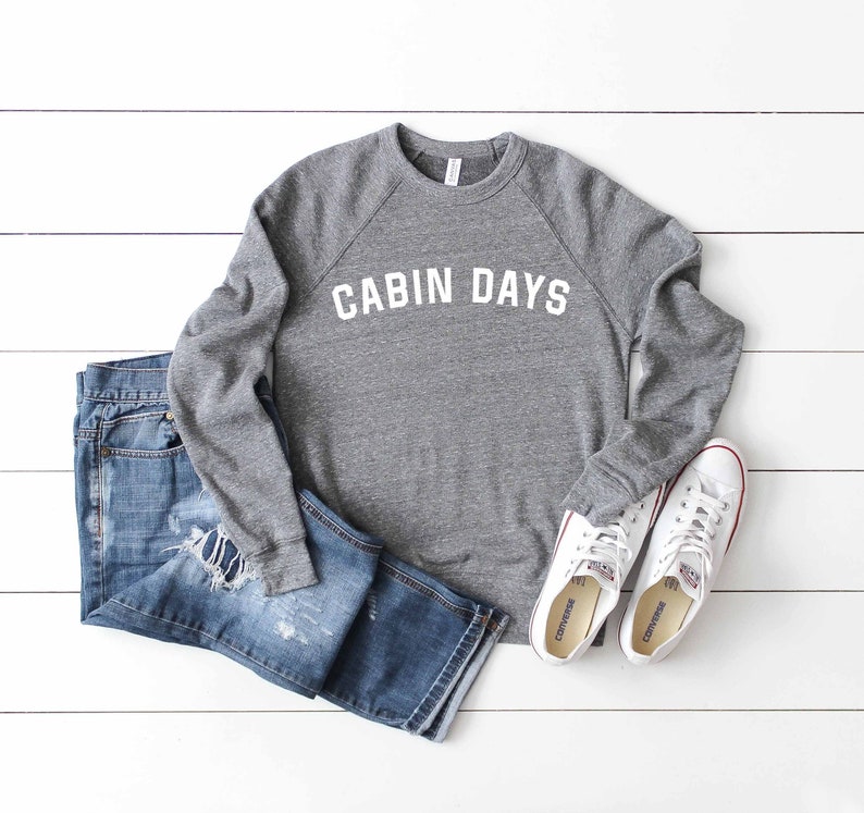 May include: A gray sweatshirt with the text "CABIN DAYS" in white lettering. The sweatshirt is paired with blue jeans and white sneakers.