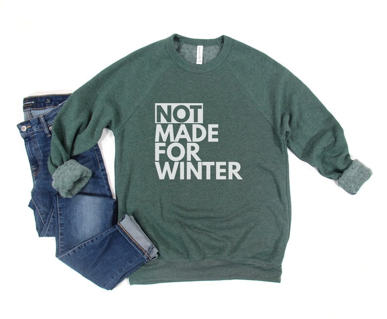 May include: A green sweatshirt with the text "NOT MADE FOR WINTER" in white, paired with blue jeans. The sweatshirt has rolled-up sleeves. The jeans are folded at the ankles.