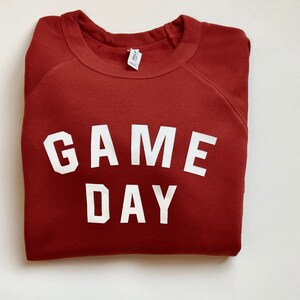 Puede incluir: Una sudadera color marr&oacute;n rojizo con el texto "GAME DAY" impreso en blanco.