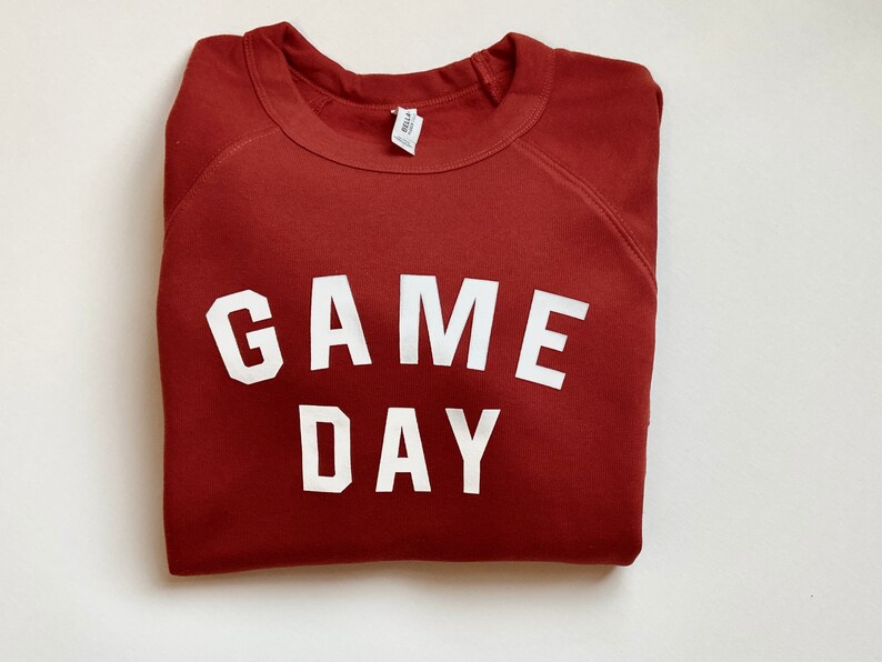 Puede incluir: Una sudadera color marr&oacute;n rojizo con el texto "GAME DAY" impreso en blanco.