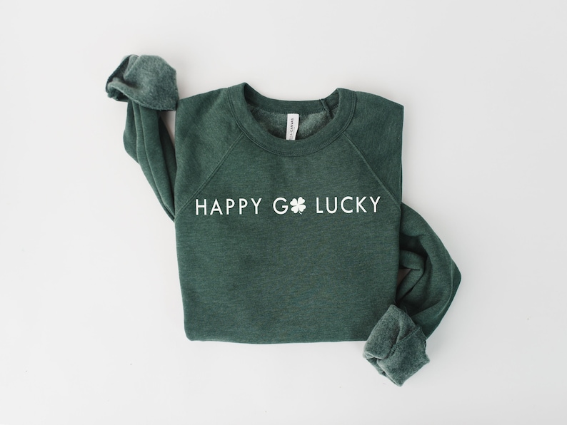 May include: A dark green sweatshirt with the words "HAPPY GO LUCKY" printed in white. A white shamrock graphic is between the words "GO" and "LUCKY". The long sleeves are cuffed. The sweatshirt is on a white background.