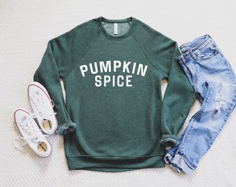 May include: A green sweatshirt with the text "Pumpkin Spice" in white letters. The sweatshirt is laid out on a white surface with a pair of white sneakers and blue jeans.
