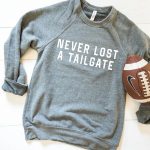 May include: Gray sweatshirt with the text "Never Lost A Tailgate" in white lettering. A brown and white football is lying on the right side of the sweatshirt.