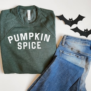 May include: A green sweatshirt with the text "PUMPKIN SPICE" in white letters. The sweatshirt is folded and laid on a white surface next to a pair of blue jeans and two black paper bats.